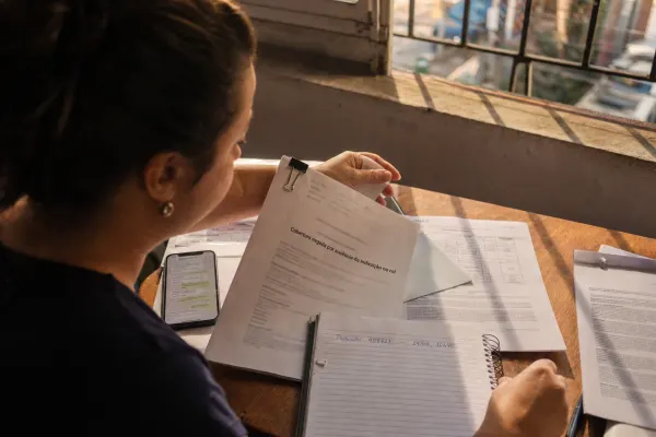 Mulher sentada à mesa perto da janela segura documento com o texto “Cobertura negada por ausência de indicação no rol”, ao lado de celular, papéis e caderno com “Protocolo 458721 - 14/08, 16h40”.