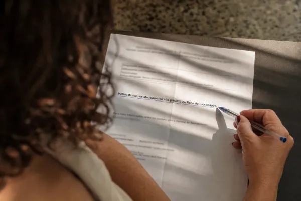 Mulher vista de costas sublinha com caneta em um documento impresso a frase: Motivo da recusa: Medicamento não previsto no Rol e de uso off-label.
