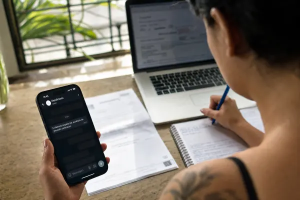 Pessoa sentada à mesa segura celular com mensagem “Cobertura negada por ausência de previsão contratual”, enquanto escreve em caderno diante de notebook e papéis.