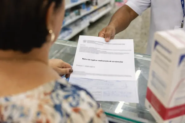 Mulher no balcão de uma farmácia recebe de um atendente um documento com o texto “Motivo da negativa: medicamento de uso domiciliar”.