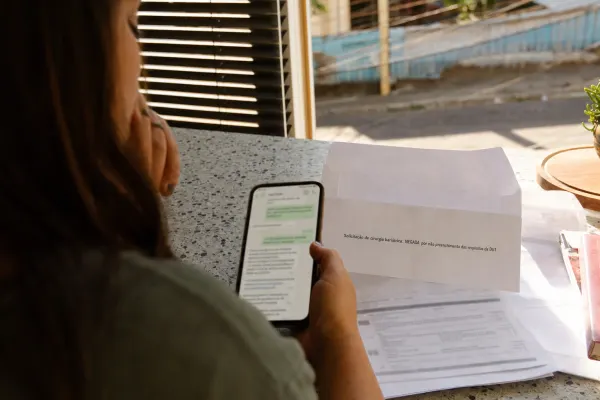 Pessoa sentada à mesa, olhando o celular ao lado de papéis e de uma carta aberta com o texto: “Solicitação de cirurgia bariátrica: NEGADA por não preenchimento dos requisitos da DUT”.